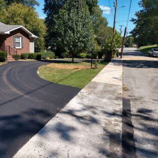Suburban driveway beside a house, with a concrete sidewalk and trees casting shadows along the road.