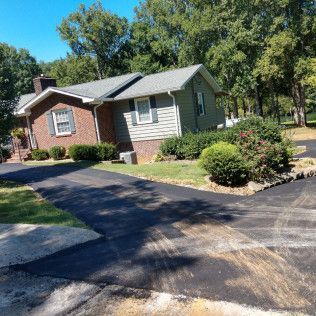 Suburban house with brick and gray siding, black driveway, and trees in the yard under a clear blue sky