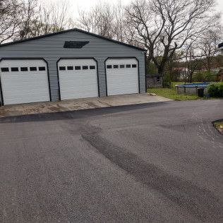 Gray three-bay garage with white doors beside a paved driveway and leafless trees