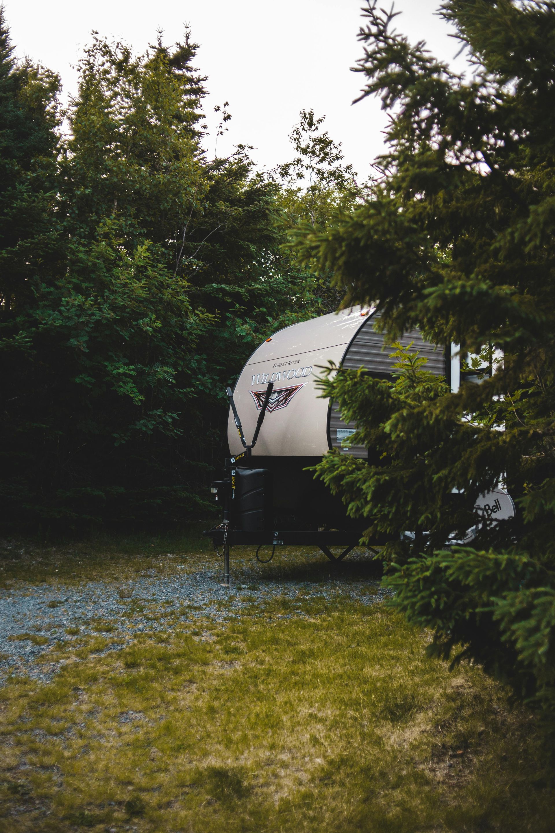 Small camper with a curved roof, surrounded by trees on a gravel lot.