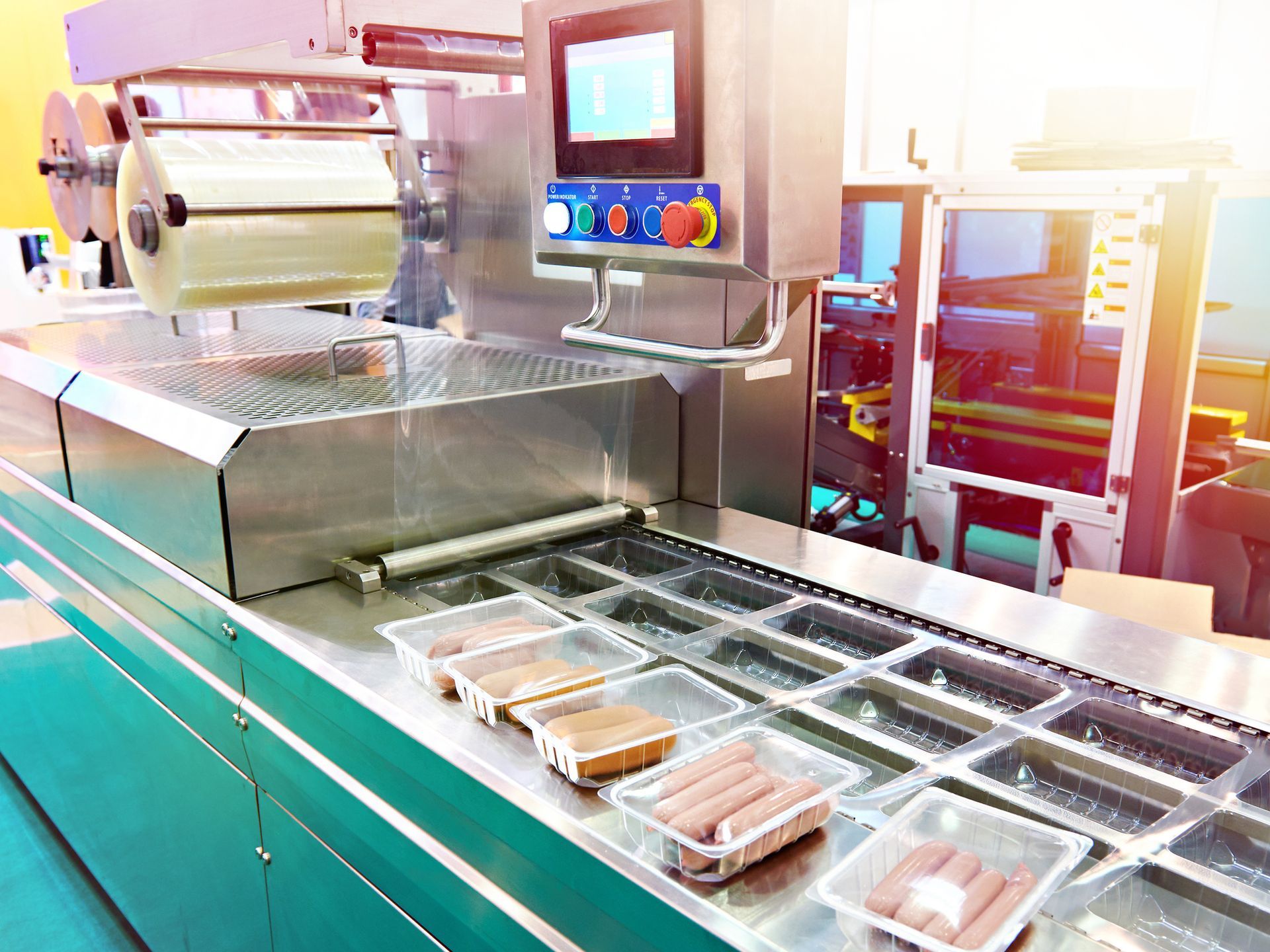 Packaging machine sealing trays of food, likely sausages, in a factory setting.
