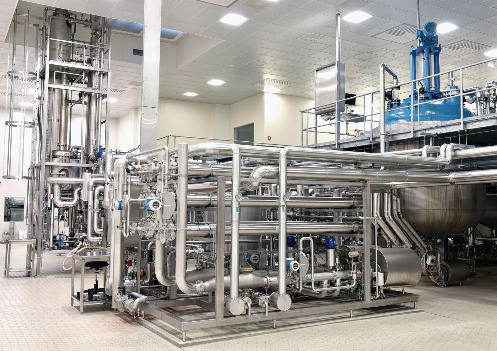 Industrial stainless steel processing equipment in a cleanroom environment. Pipes, tanks, and machinery are visible.