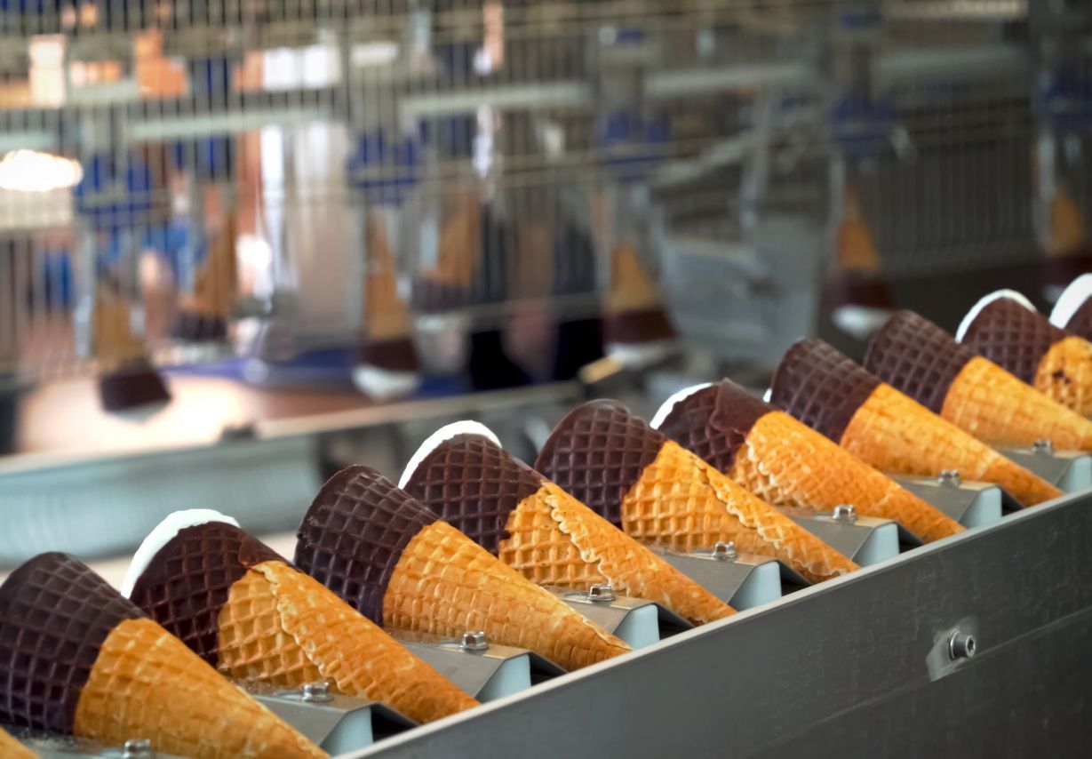 Ice cream cones dipped in dark chocolate move along a metal industrial conveyor belt in a factory.