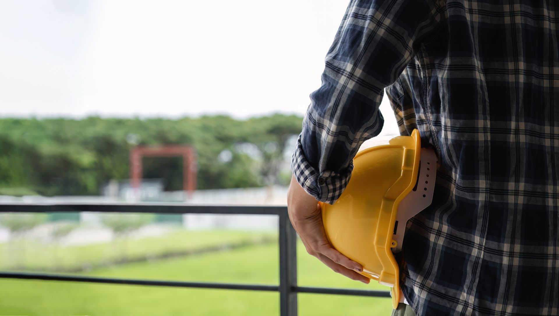 A person in a plaid shirt holds a yellow hard hat at their side, standing on a balcony overlooking a green field.