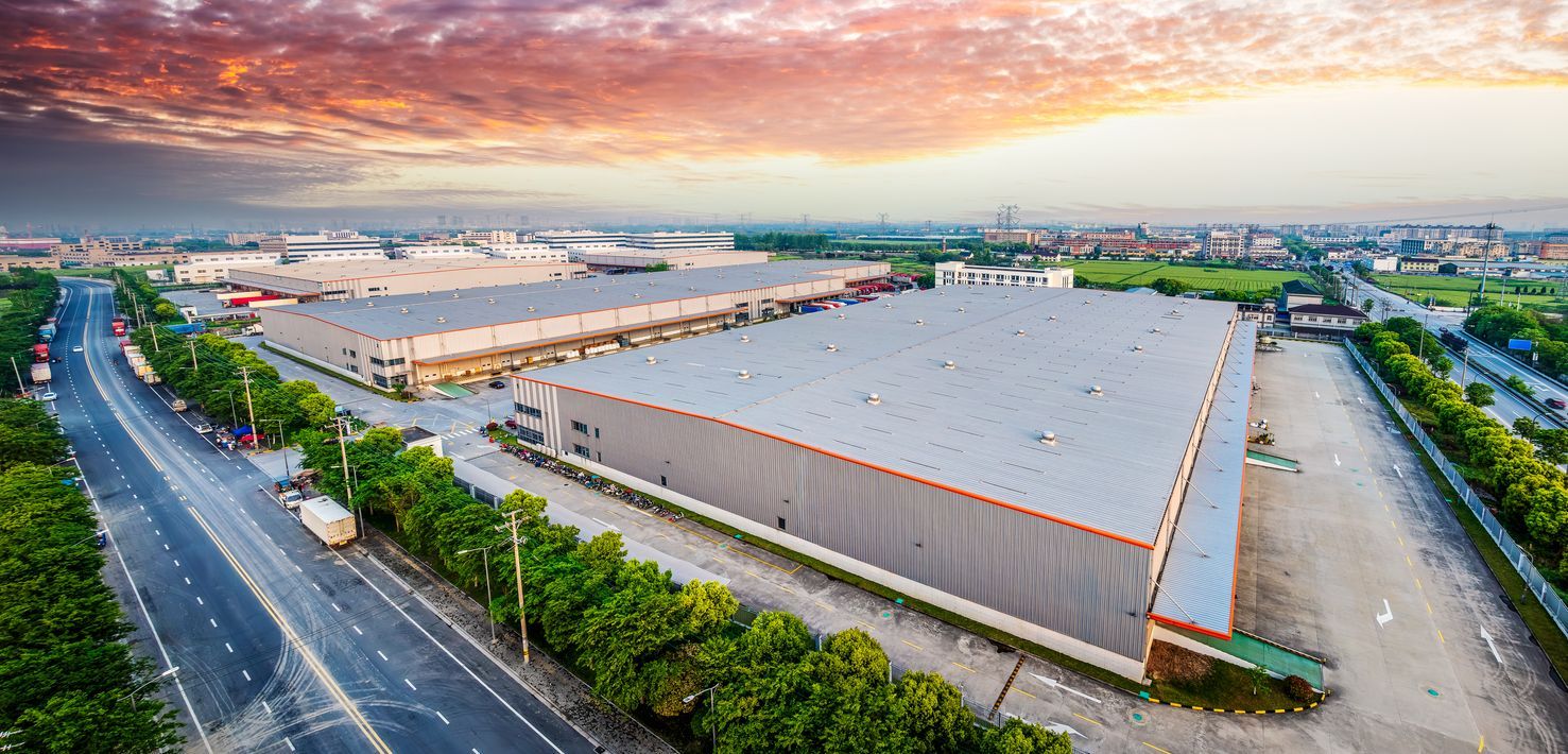 Aerial view of warehouses with a road and trees against a sunset sky.