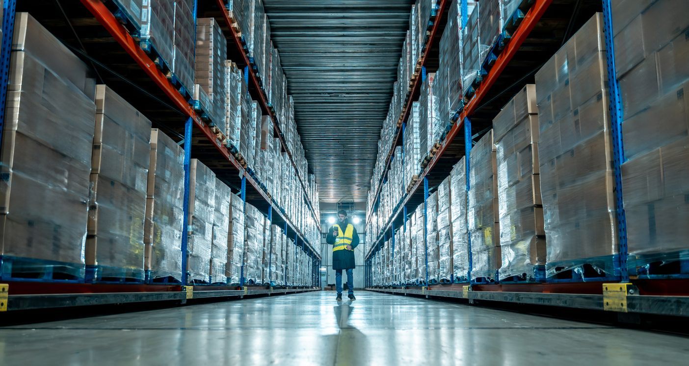 A person in a high-visibility vest walks down the aisle of a vast warehouse filled with tall shelves of boxed inventory.