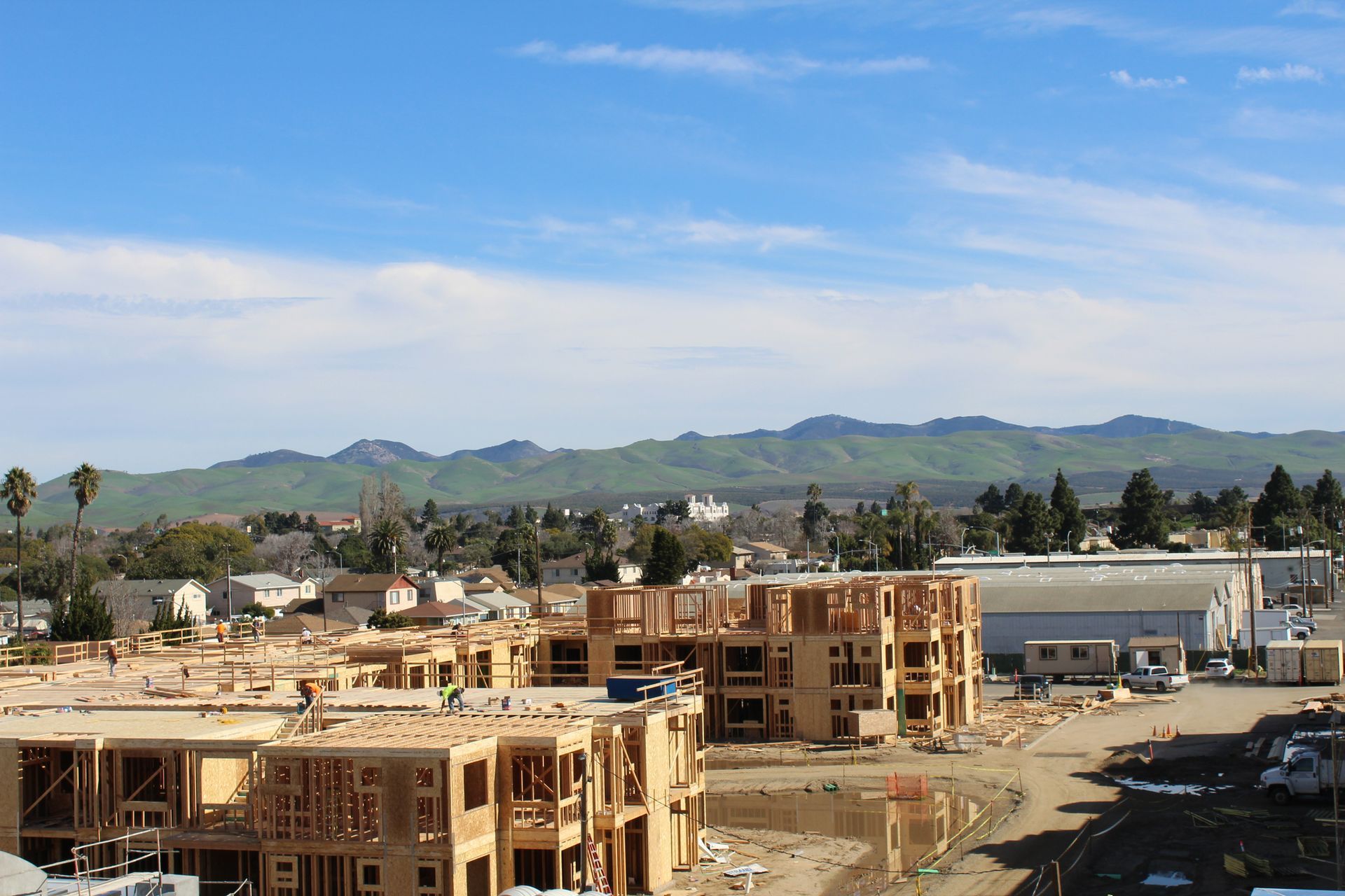Construction of multi-story wood-frame buildings under a blue sky with a mountain range in the background.