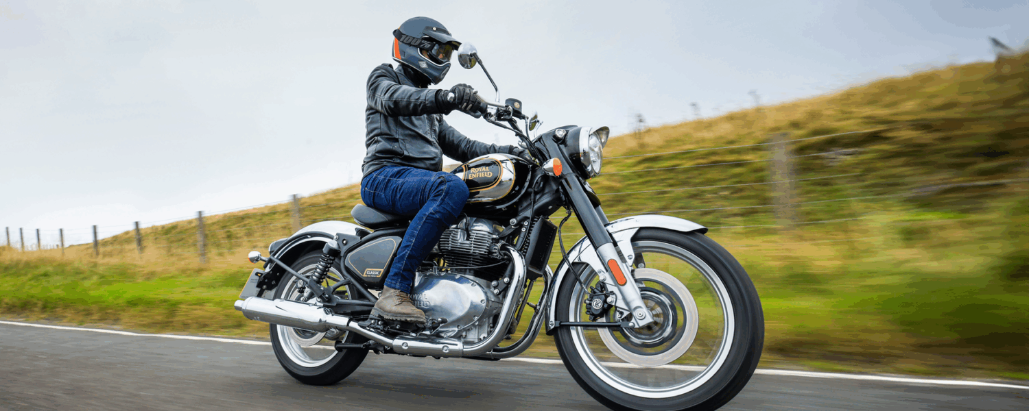 Motorcyclist riding a chrome bike on a road, with blurred grassy hills in the background.