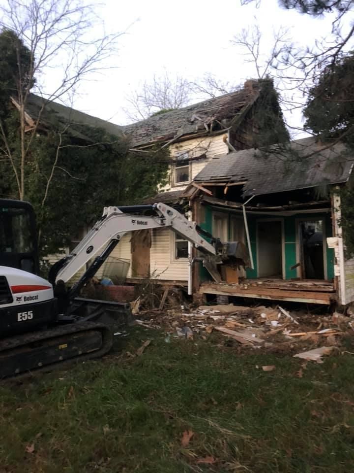 A small excavator demolishing a weathered, two-story house with a damaged roof and open walls.