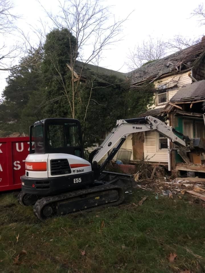 Bobcat excavator at a house demolition site, pulling down a building. Red dumpster on the left.