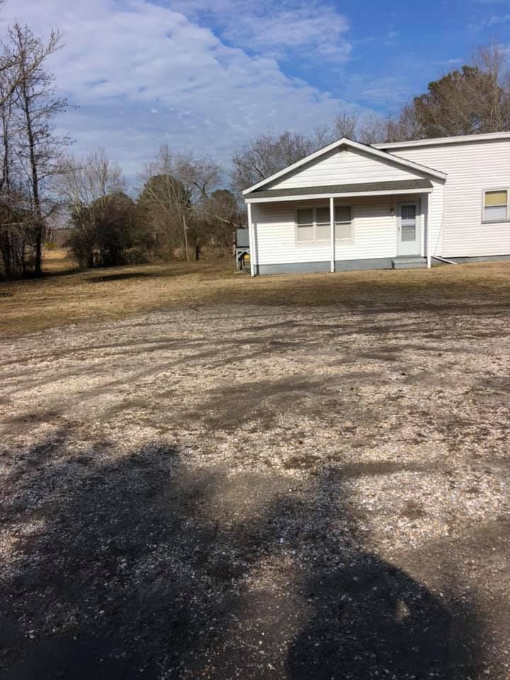 White building with a porch and windows on a sunny day; dirt lot in front, trees and blue sky.