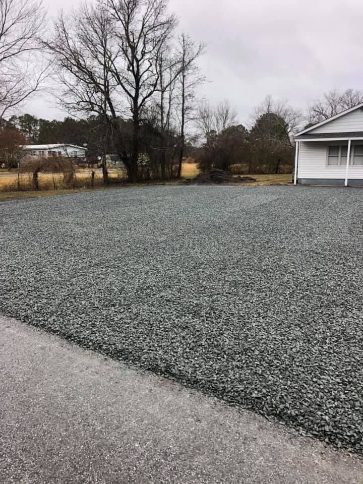 Gravel driveway next to asphalt road in front of a white house on a cloudy day. Bare trees in the background.