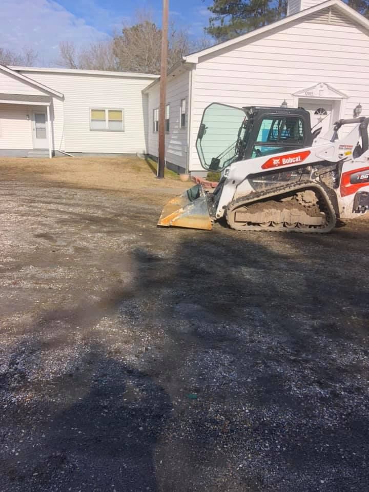 Bobcat skid-steer with a leveling attachment is working in a paved area, next to a white building.