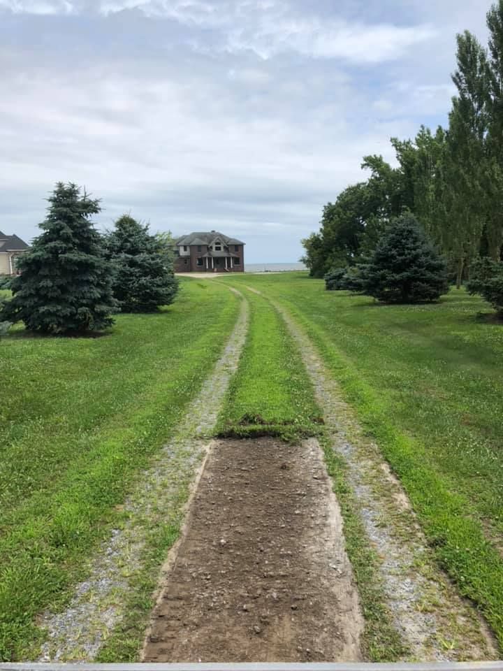 Gravel driveway leading toward a house on a grassy lawn with trees, under a cloudy sky.