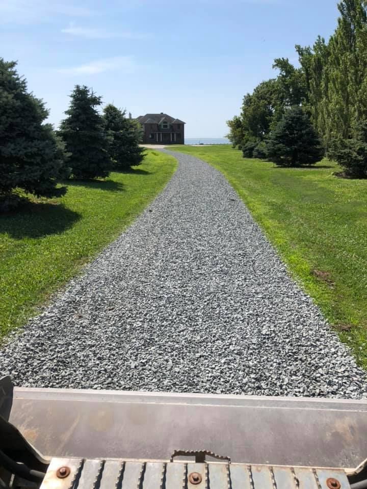 Gravel path leading to a building near water, flanked by green grass and trees. Blue sky overhead.
