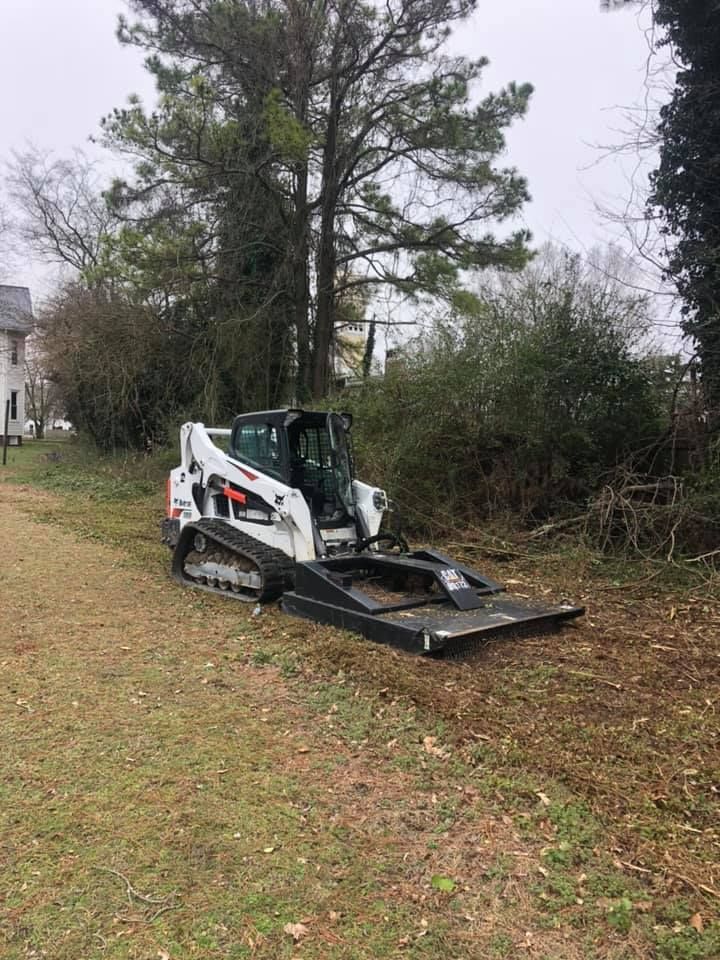 Bobcat skid steer with brush cutter attachment on a grassy area, clearing overgrown vegetation near trees.