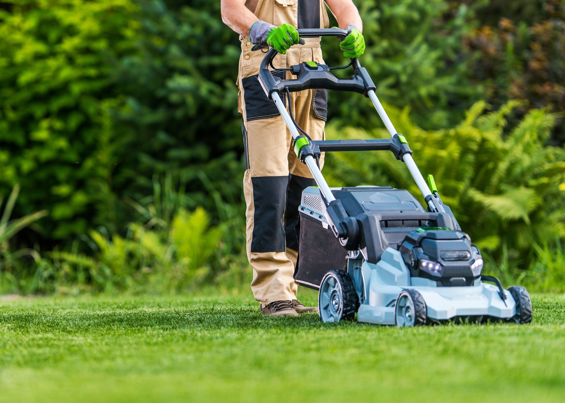 Person mowing a green lawn with a gray and black lawnmower in a garden setting.