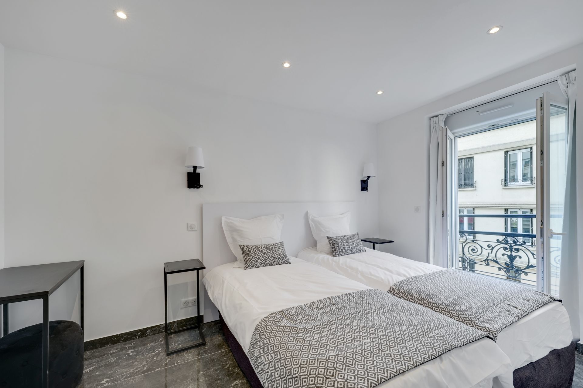 Bedroom with two beds, white walls, a balcony, and a black desk.