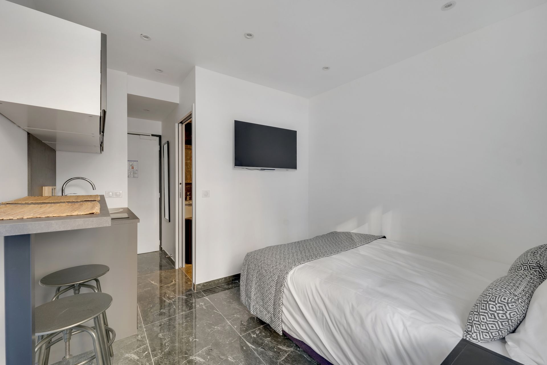 Small, modern studio with bed, TV, kitchenette, and bathroom entrance.