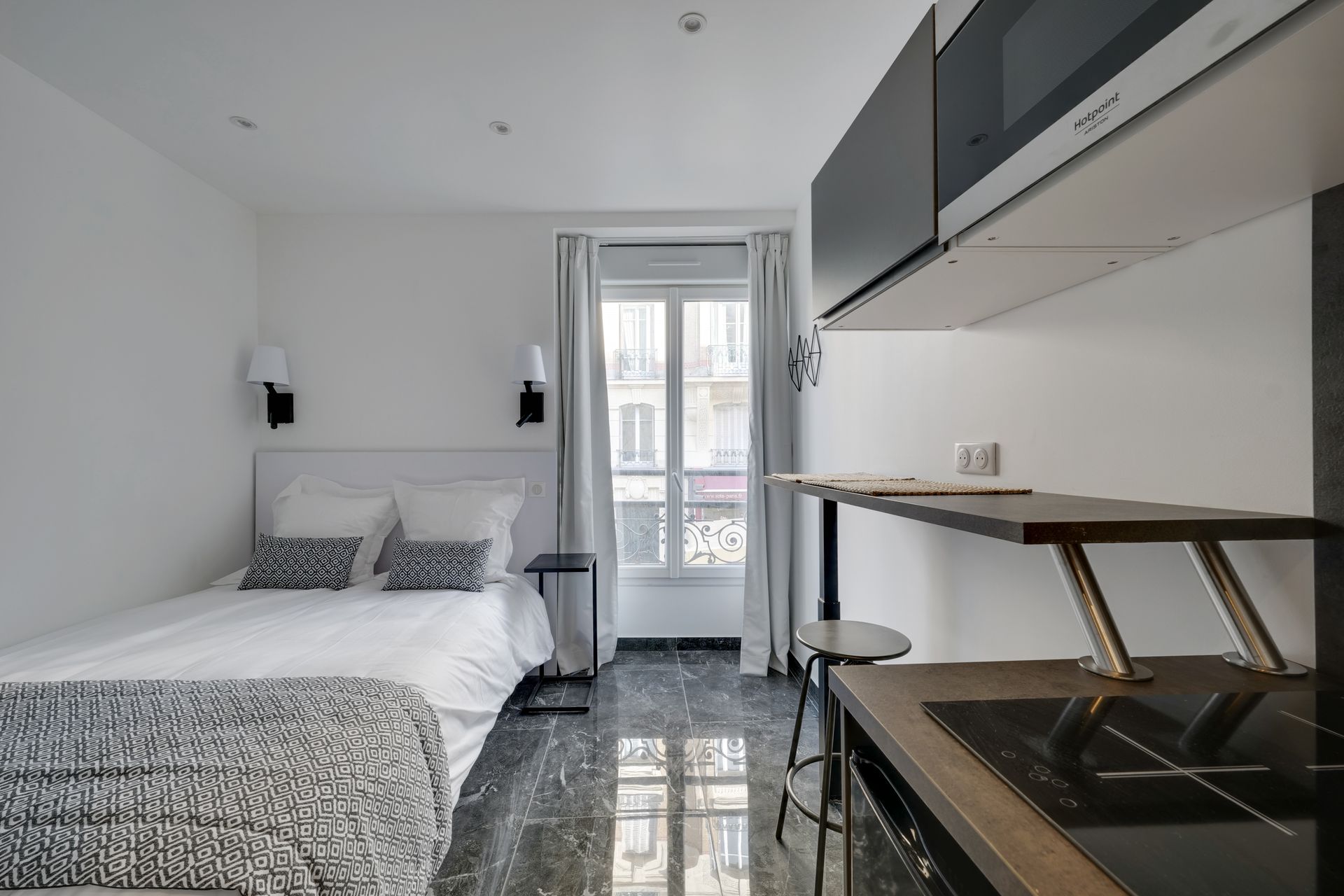 Small, modern studio with bed, kitchenette, and window looking out to a street.