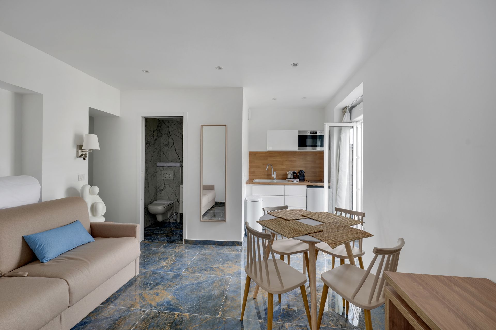 Bright apartment interior with living, dining, and kitchen areas. Features include a sofa bed, table, and a tiled floor.