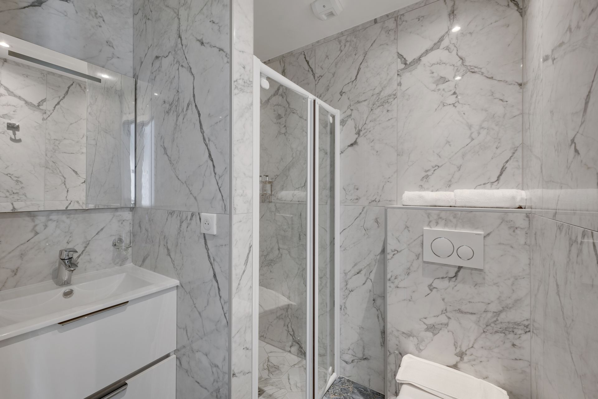 Modern bathroom with marble-patterned walls, glass shower, white sink, toilet, and mirror.