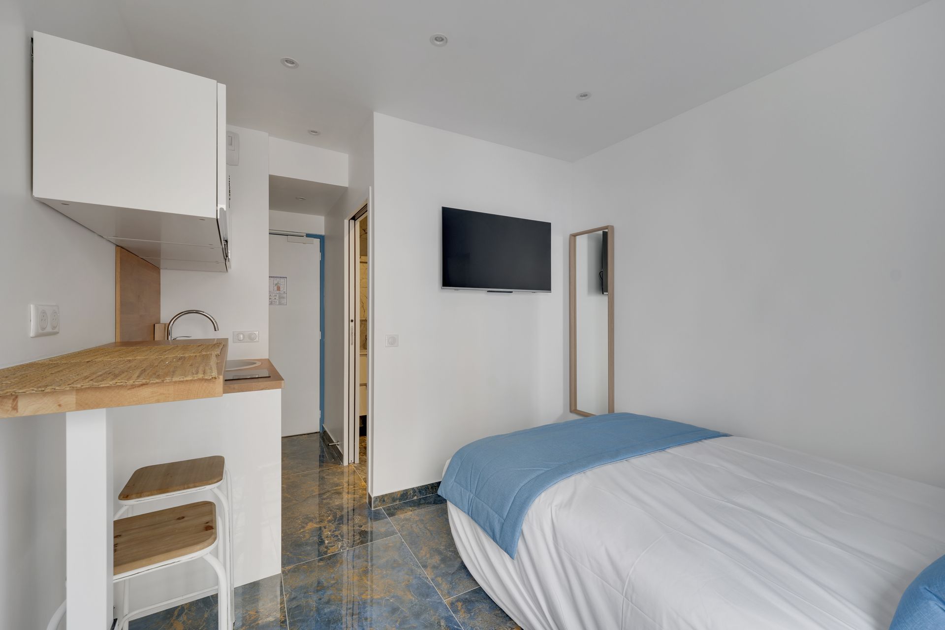 Small, bright studio with a bed, desk, TV, and long mirror; white walls.