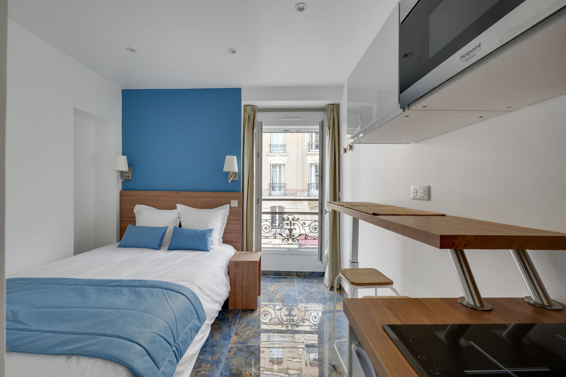 Small studio with bed, blue wall, small kitchen area, and a window.