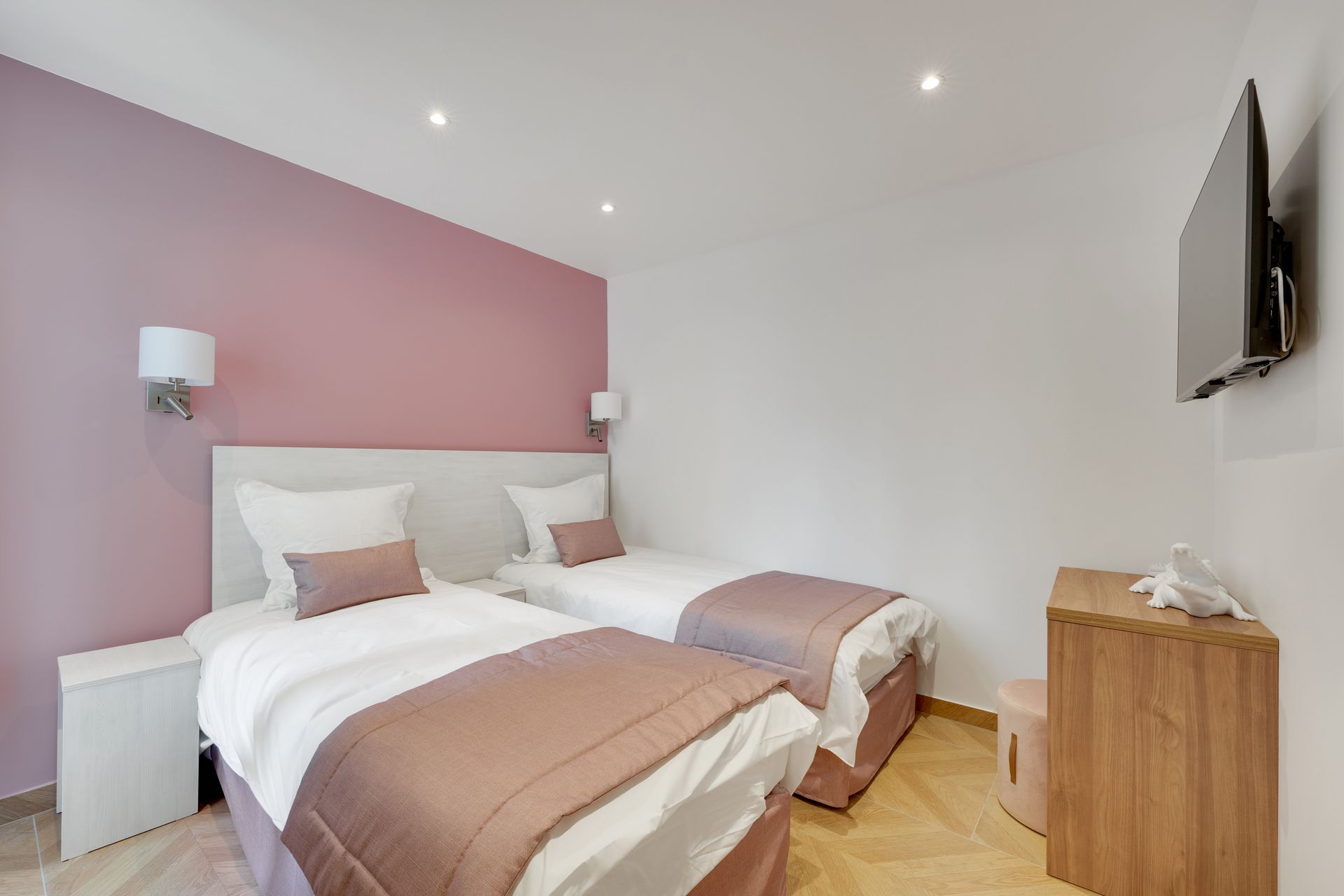 Twin beds in a hotel room, pink bedding, white walls, wood nightstand, TV.