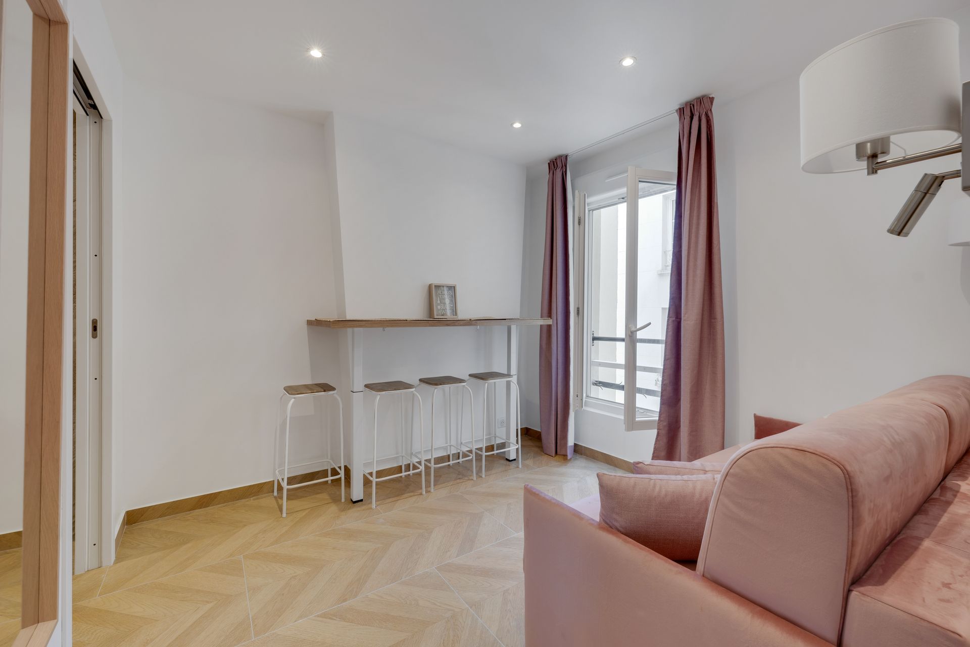 A small, modern living space with a pink sofa bed, bar-style seating, and a window with pink curtains.