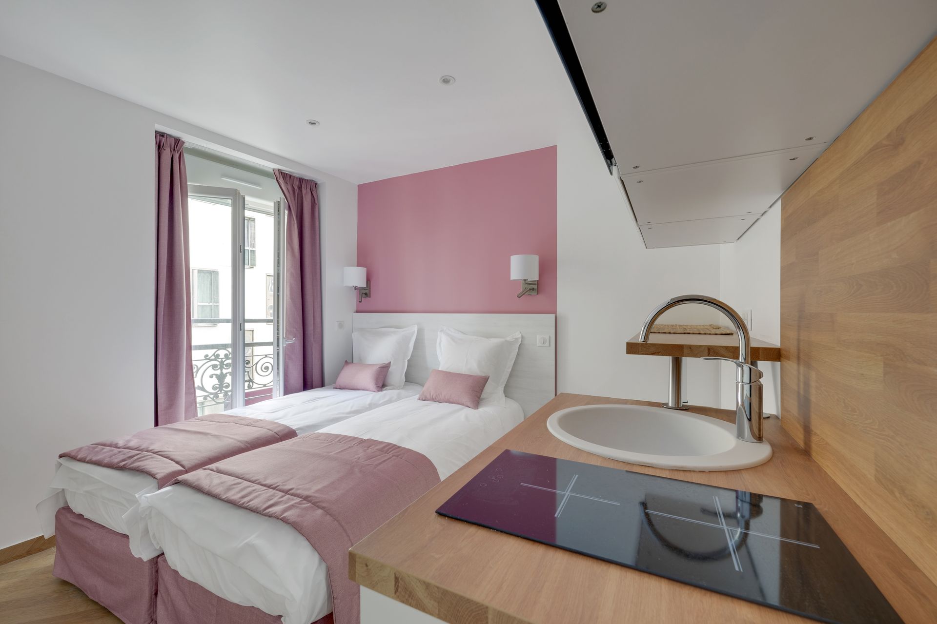 Small studio with two twin beds, pink accent wall, and a small kitchenette.
