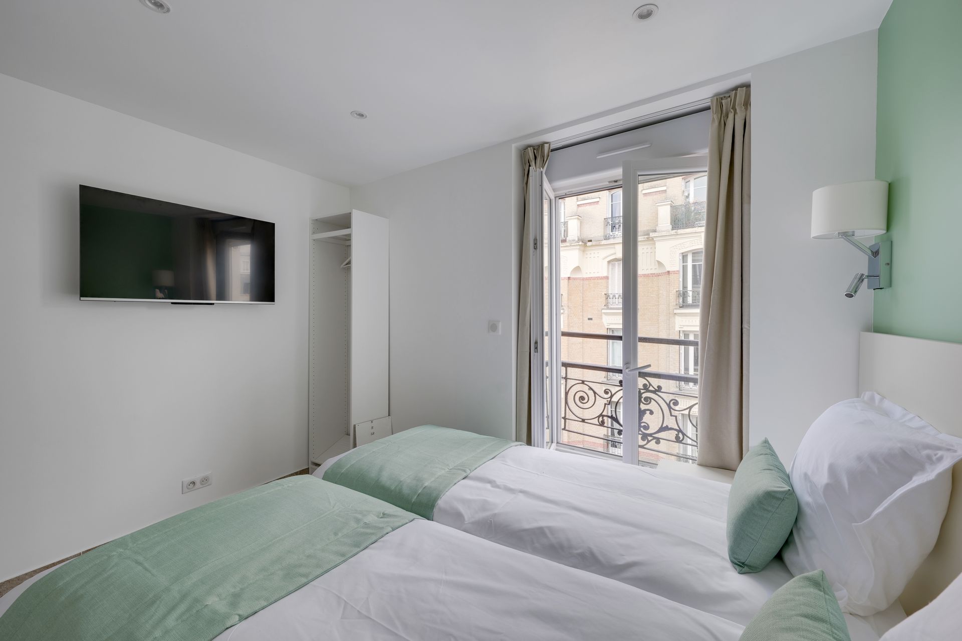 A bright studio with twin beds, balcony, TV, and wardrobe; white and mint green decor.