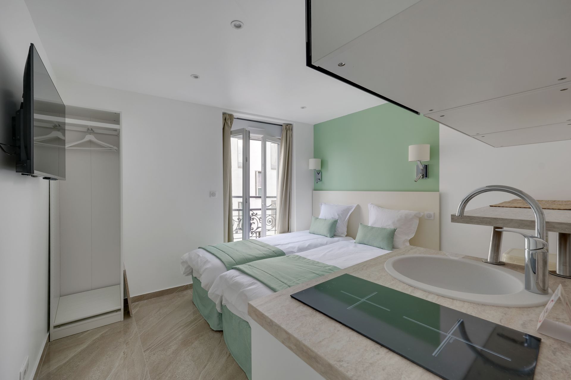 Small, bright studio with twin beds, kitchenette, and television. Soft green and white color scheme.