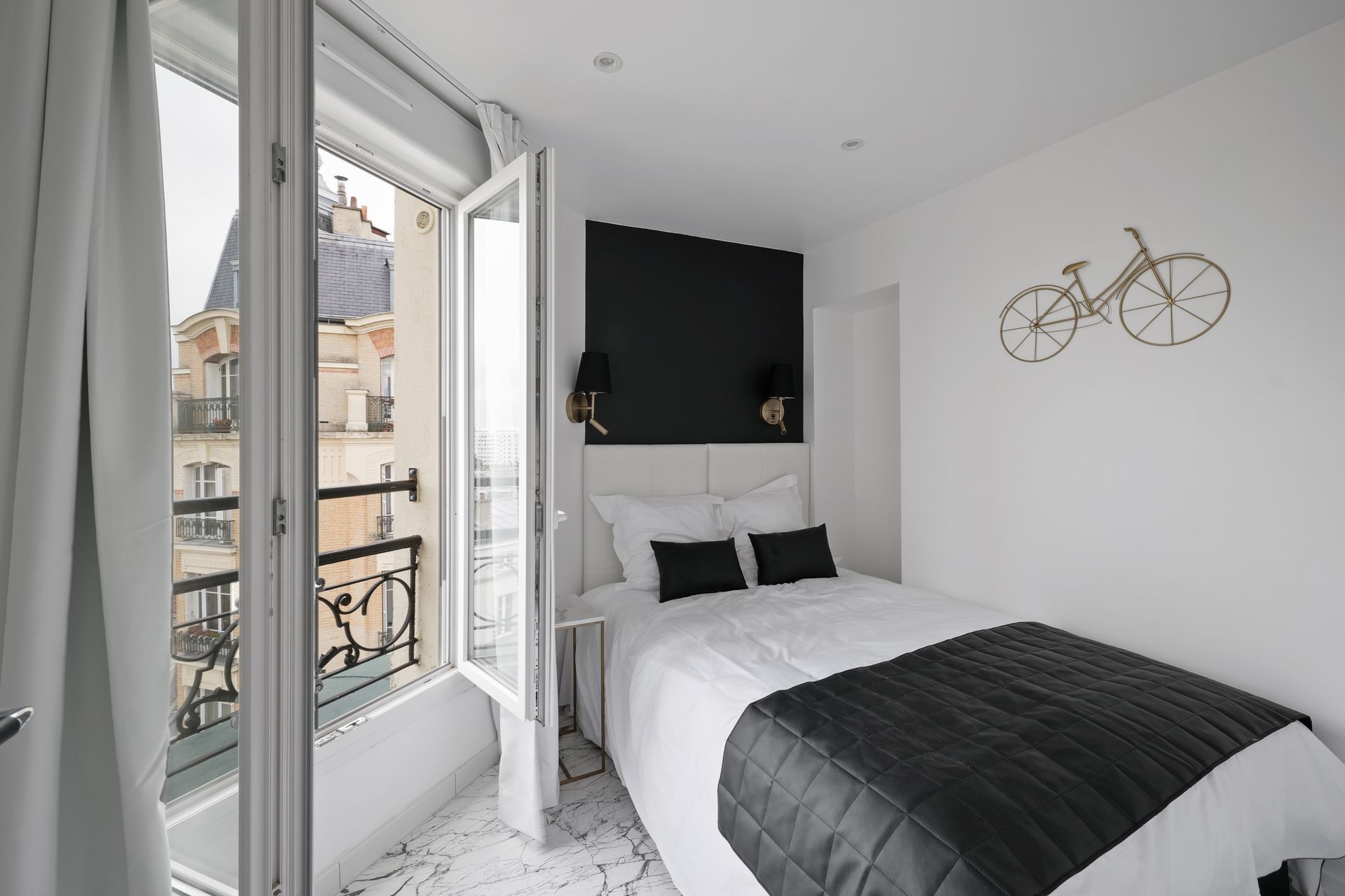 Small, white bedroom with open window, a bed with black and white bedding, and a bicycle wall art.