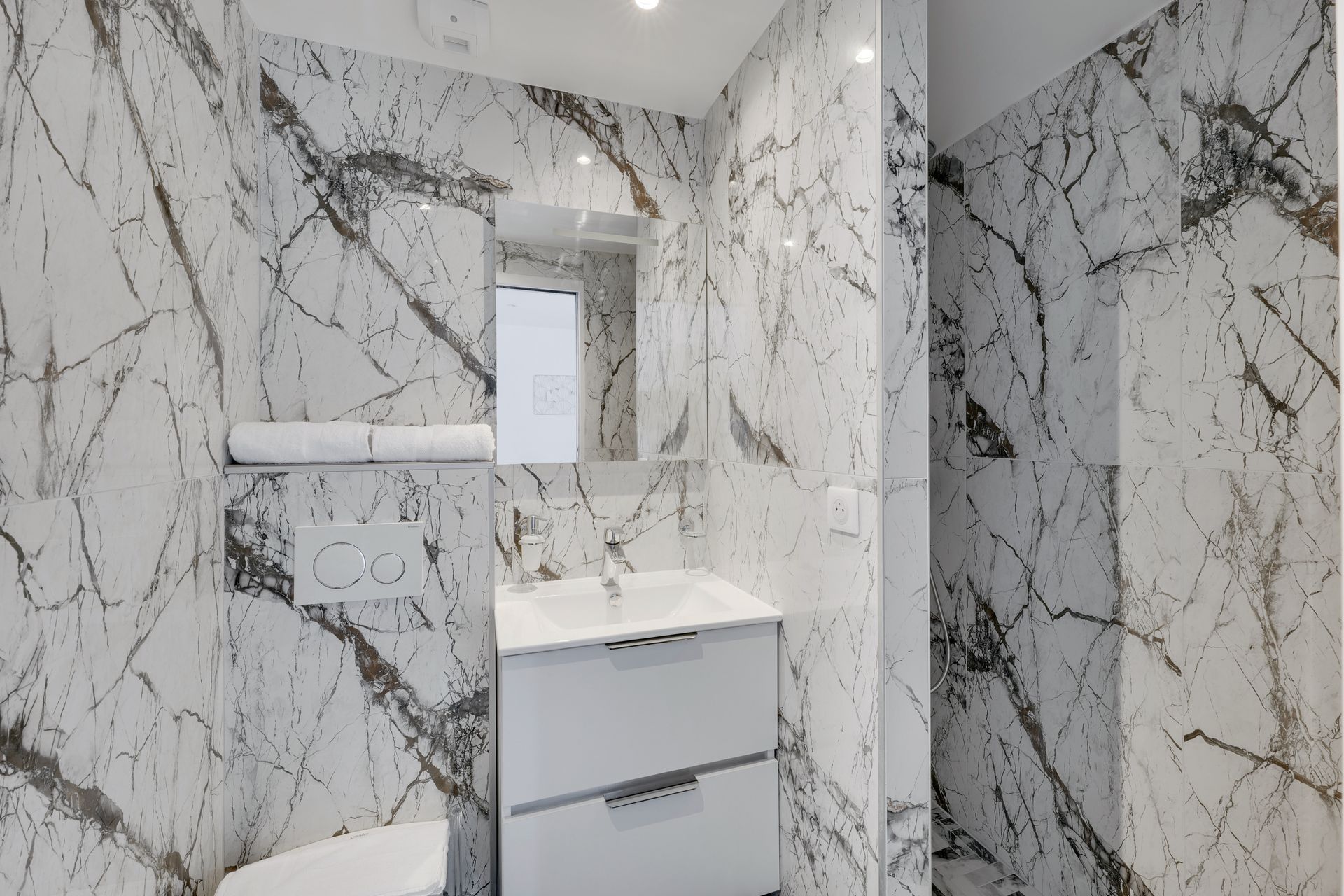 Bathroom with white marble-look tiles, white vanity, and mirror. A roll of towels is mounted above the toilet.
