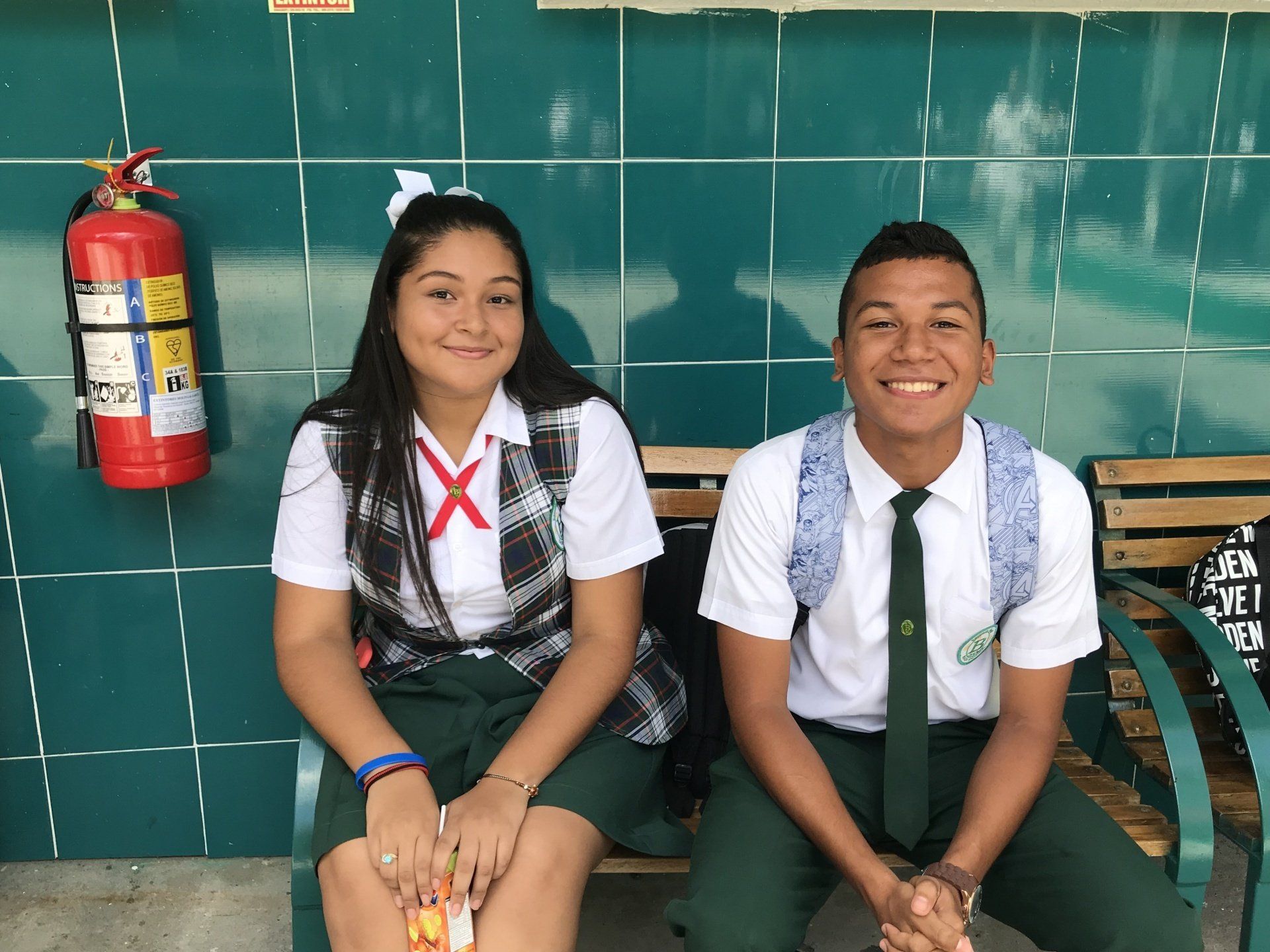 A boy and a girl are sitting on a bench in front of a fire extinguisher