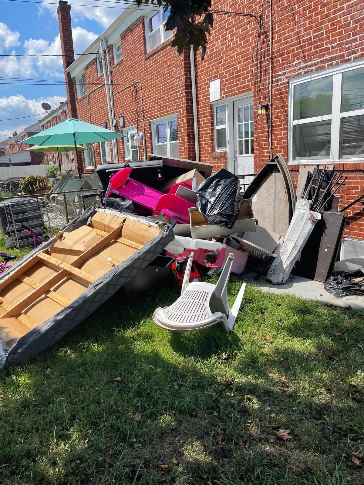 Pile of trash in a yard: wood, plastic, and other debris. Red brick building and green grass in background.
