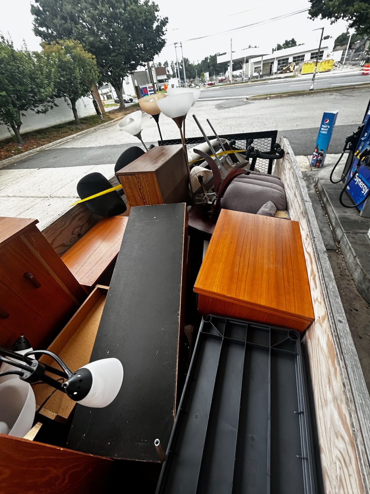 Truck bed overflowing with furniture at a gas station. Wooden cabinets, a lamp, and plastic bins.