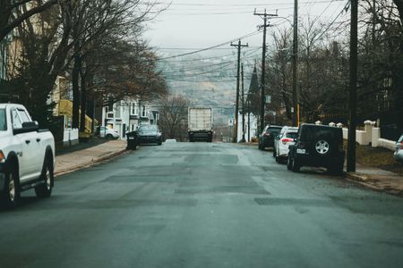 Street with parked cars, a semi-truck, houses, and power lines on a cloudy day.