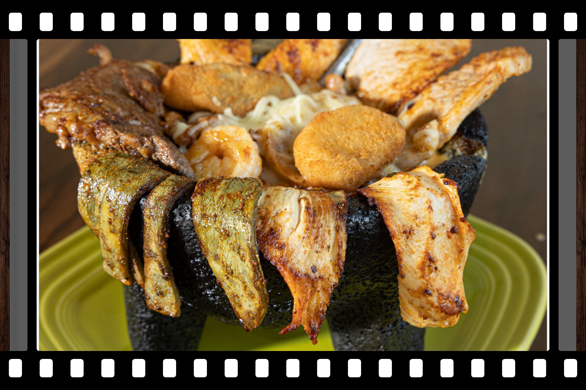 Molcajete dish: assorted meats and vegetables in a volcanic stone bowl, on a green plate.