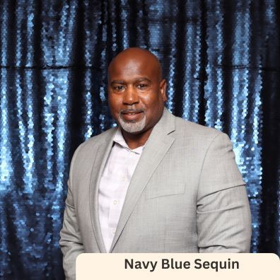 A man in a suit is standing in front of a navy blue sequin backdrop.