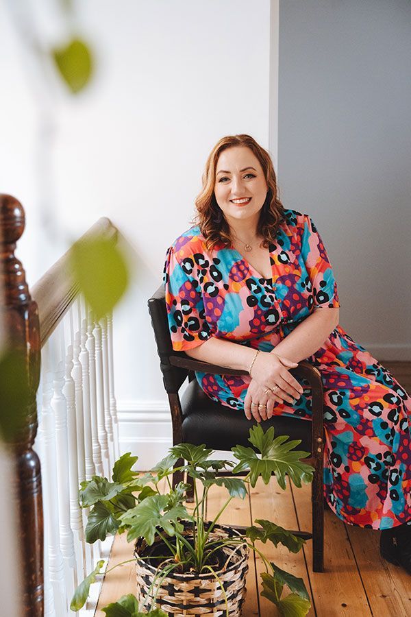 A woman in a colourful dress is sitting in a chair next to a potted plant.
