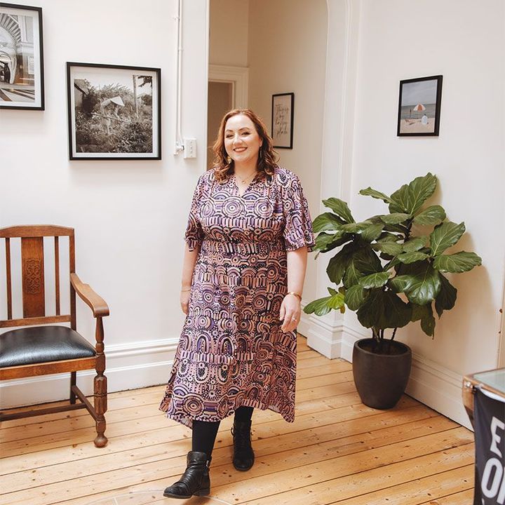 A woman in a dress is standing in a room next to a chair and a plant.