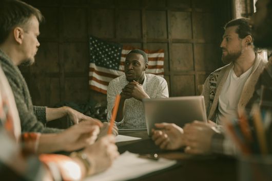 Group of people in a meeting. A man in the center looks at the others. An American flag is behind them.