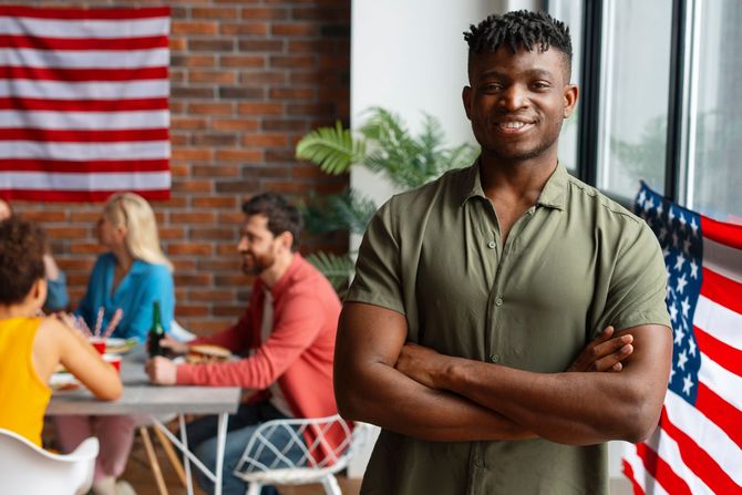 Man with crossed arms smiles; group of people seated at a table with an American flag.