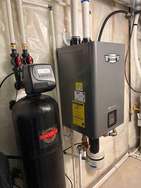 Water softener and gray HVAC control box in a basement with insulated walls and exposed pipes.