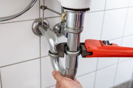 Hand tightening a chrome sink pipe with a red pipe wrench under a bathroom sink