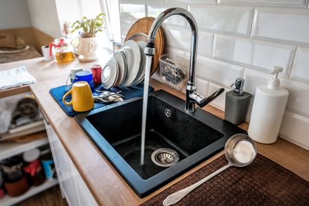 Kitchen sink with running faucet, black basin, blue mat, yellow mug, and white tiled backsplash