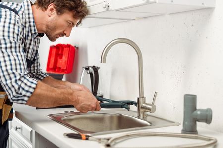 Man repairing a kitchen faucet beside a sink with tools on the counter