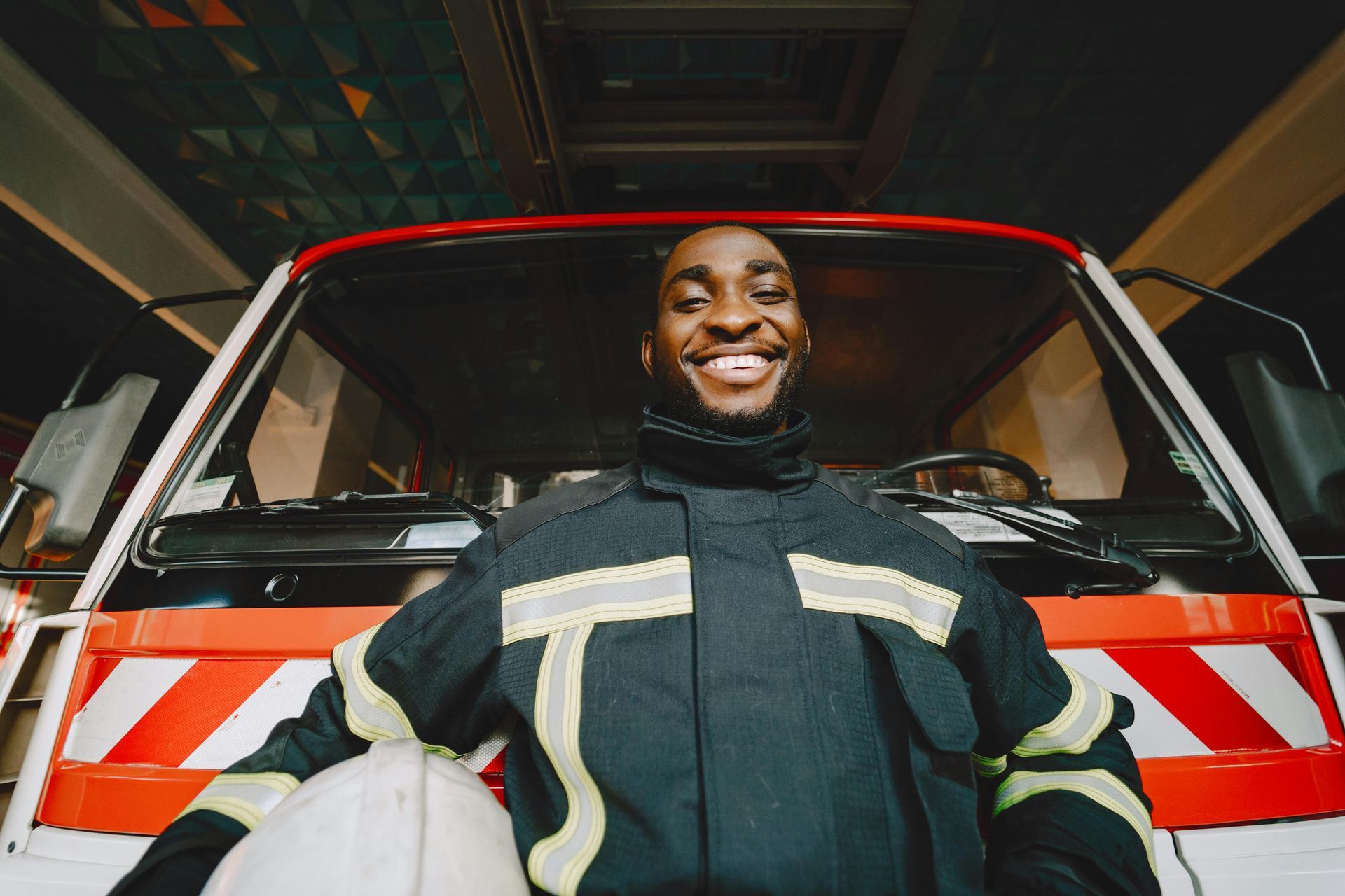 Smiling Black firefighter standing in front of a fire truck, holding a helmet.
